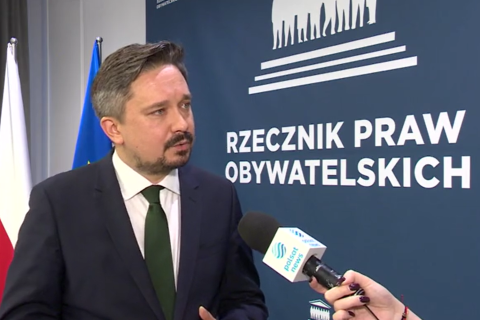 RPO Marcin Wiącek wypowiada się przed mikrofonem, w tle granatowa ścianka z logotypem RPO oraz flagi Polski i Unii Europejskiej