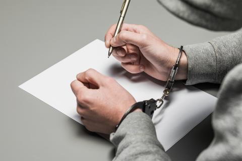 Mężczyzna w kajdankach z długopisem w dłoni pochyla się nad kartką papieru