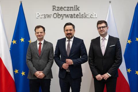 Adam Krzywoń, Marcin Wiącek i Valeri Vachev pozują do zdjęcia na tle flag Polski i Unii Europejskiej oraz napisu "Rzecznik Praw Obywatelskich"