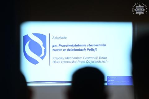 Ekran projektora z napisem "Szpolenie pn. Przeciwdziałanie stosowaniu tortur w działaniach Policji, Krajowy Mechanizm Prewencji Tortur BRPO"