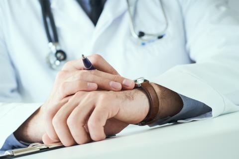 Zbliżenie na położone na blacie biurka dłonie mężczyzny w białym fartuchu i z przewieszonym przez szyję stetoskopem. Mężczyzna sprawdza na zegarku godzinę.