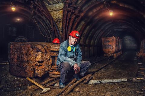 Mężczyzna w kasku ochronnym, z maską przeciwpyłową zawieszoną na szyi, siedzi w szybie kopalni