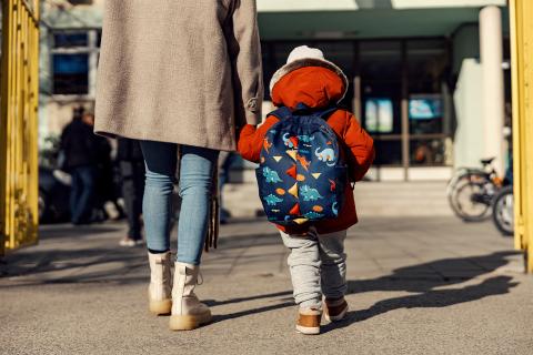 Widok od tyłu dorosłej osoby prowadzącej za rękę kilkuletnie dziecko z tornistrem w stronę drzwi wejściowych budynku