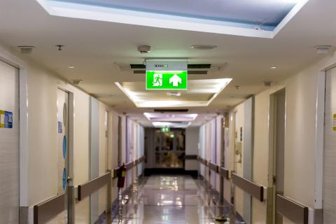 Widok długiego korytarza w szpitalu. Na suficie podwieszone są podświetlone na zielono znaki wskazujące drogę do wyjścia z budynku.