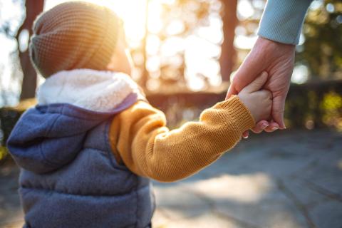 Kilkuletnie dziecko w czapce idzie na spacerze w słoneczny dzień trzymając się za ręce z osoba dorosłą