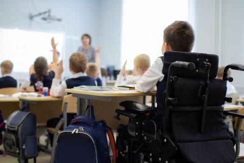 Dziecko siedzące na wózku przy ławce w sali szkolnej pośród innych dzieci, które słuchają nauczyciela