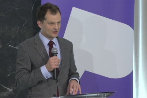 Adam Krzywoń stoi przy podium i wypowiada się do mikrofonu
