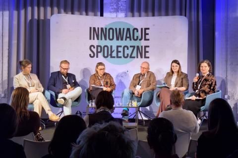 Sześć osób siedzi na scenie na krzesłach, jedna z nich mówi do mikrofonu, rozmowie przysłuchują się osoby siedzące na widowni