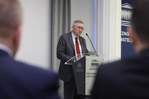 Stanisław Trociuk wypowiada się stojąc przy mównicy w sali konferencyjnej. Na pierwszym planie widać sylwetki osób siedzących na widowni