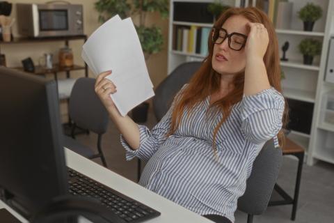 kobieta w ciąży siedzi przy komputerze trzymając plik kartek  