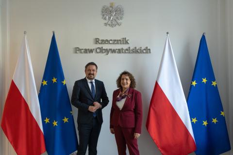 RPO Marcin Wiącek i Melinda Simmons uśmiechają się pozując do zdjęcia na tle flag Polski i Unii Europejskiej oraz godła Polski i napisu "Rzecznik Praw Obywatelskich"