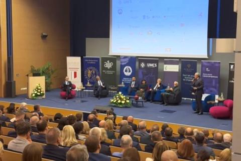 Siedem osób na scenie dużej auli wykładowej na uniwersytecie, na widowni licznie zgromadzona publiczność