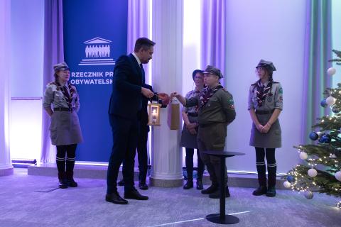 Marcin Wiącek przyjmuje od młodego harcerza lampion z zapaloną świecą. Przygląda się temu trzech innych harcerzy. Obok stoi udekorowana choinka
