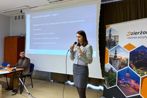 Osoba stoi i mówi do mikrofonu gestykulując, w tle na projektorze widoczny slajd prezentacji z tekstem "Dostępność w pigułce"