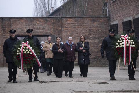 osoby stoją trzymajac wieńce na tle muru 
