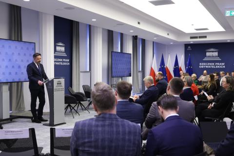 Kilkadziesiąt osób siedzi na sali konferencyjnej i słucha wystąpienia osoby mówiącej za mównicą na środku