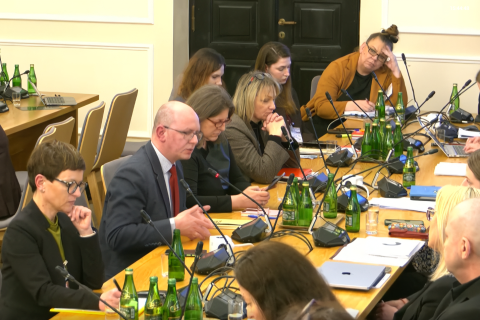 osoby siedzą w sali, jedna mówi do mikrofonu  