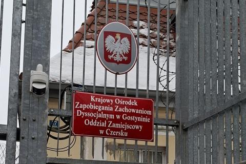 Tablice z godłem Polski i napisem "Krajowy Ośrodek Zapobiegania Zachowaniom Dyssocjalnym w Gostyninie - Oddział Zamiejscowy w Czersku" na płocie z drutem kolczastym. Obok tabliczki wisi kamera monitoringu.