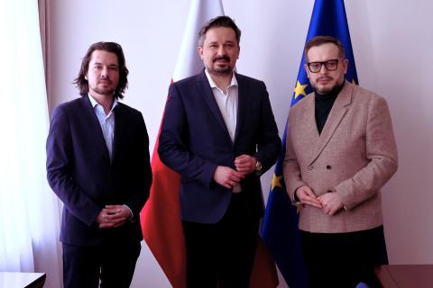 Trzy osoby pozujż do wspólnego zdjęcia na stojąco na tle flag Polski i UE