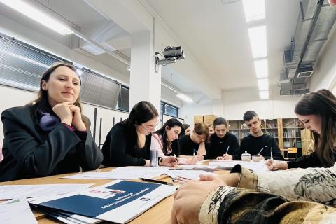 Kilka osób siedzi przy dużym stole i wypełnia dokumenty rozmawiając ze sobą