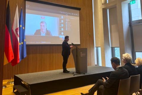 Sala konferencyjna z mównicą przy której stoi osoba, a tle na ekranie widoczny uczestnik konferencji łączący się zdalnie