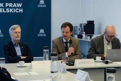 Trzy osoby siedzą przy stole w sali konferencyjnej. Jedna z osób wypowiada się trzymając mikrofon