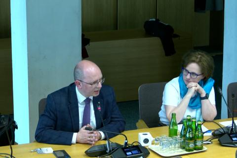 Dwie osoby siedzą za stołem, jedna mówi do mikrofonu