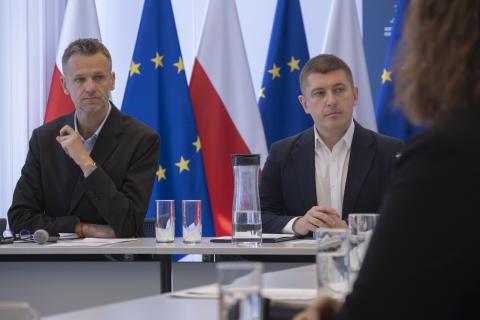 Dwie osoby siedzą przy stole na sali konferencyjnej na tle flag Polski i UE