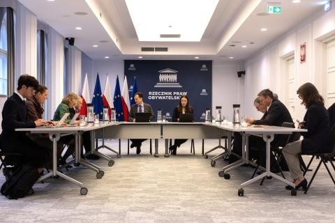 Kilkanaście osób siedzi przy stołach ustawionych w podkowę, za stołami przy ścianie rząd flag Polski i UE i duża plansza z białym napisem Rzecznik Praw Obywatelskich na granatowym tle