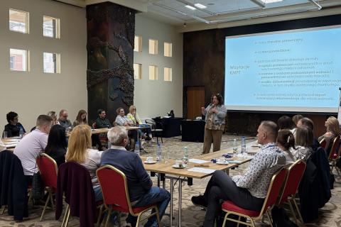 Kilkanaście osób siedzi przy stołach ustawionych w podkowę. Między stołami stoi kobieta z mikrofonem wygłaszająca prezentację, której treść wyświetla się na znajdującym się za nią dużym ekranie