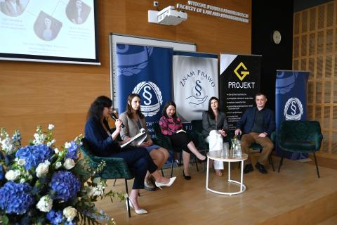 Pięć osób siedzi na krzesłach na sali wykładowej na uniwersytecie, w tle plansze z logotypami i nazwami organizatorów wydarzenia