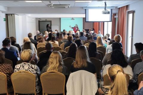 Kilkadziesiąt osób siedzi na krzesłach w sali konferencyjnej. W szczycie sali kobieta wygłasza prezentację na stojąco