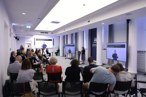 Kilkadziesiąt osób siedzi na sali konferencyjnej i słucha wystąpienia osoby mówiącej za mównicą na środku