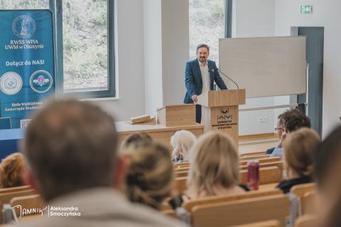 Osoba przemawia za mównicą na auli wykładowej. Na mównicy napis "Uniwersytet Warmińsko-Mazurski w Olsztynie"