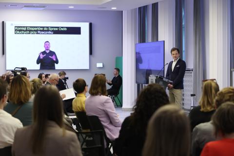 Adam Krzywoń stoi przy mównicy w sali konferencyjnej i wypowiada się w stronę siedzącej przed nim publiczności. Na ścianie jest ekran, na którym wyświetla się tłumacz języka migowego