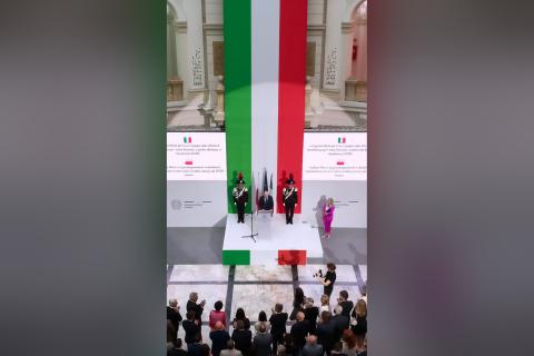 Zdjęcie z góry dużego, zabytkowego pomieszczenia z osobą przemawiającą za mównicą na podium na tle pionowo wiszącej flagi Włoch w kolorach zielonym, biąłym i czerwonym, wystąpieniu przysłuchują się zgromadzone w pomieszczeniu osoby