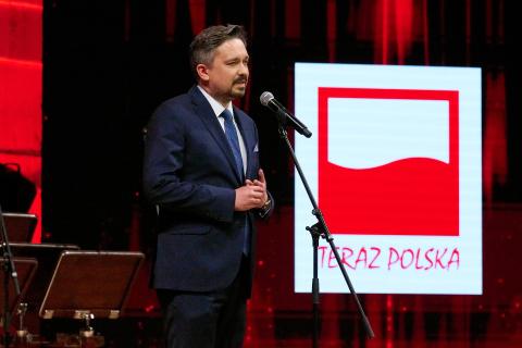 Osoba przemawia na stojąco na scenie, w tle znak graficzny z biało czerwoną flagą i napisem "Teraz Polska"