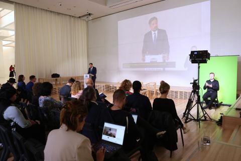 Widok sali konferencyjnej, w której siedzi kilkadziesiąt osób. Przed nimi przy mównicy stoi mężczyzna i wypowiada się, po prawej stronie na scenie na zielonym tle widoczny tłumacz PJM