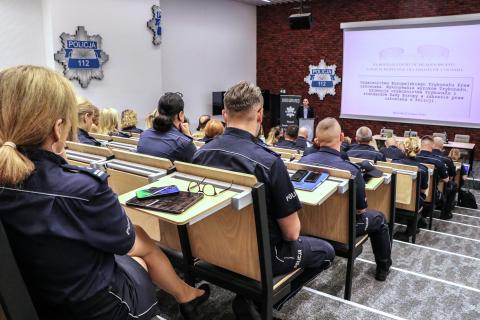 Kilkudziesięciu policjantów w mundurach słucha wystąpienia osoby mówiącej za mównicą na sali wykładowej