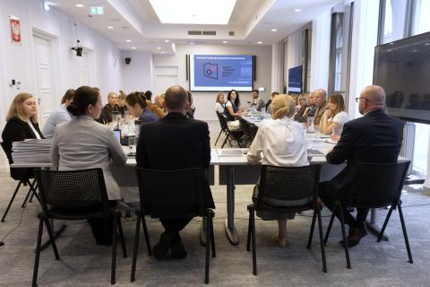 Kilkadziesiąt osób siedzi przy stołach ustawionych w podkowę na dużej sali konferencyjnej