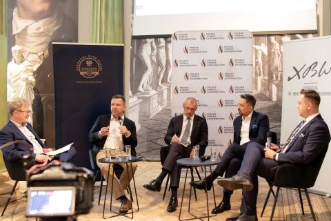 Pięć osób siedzi na krzesłach przy stolikach na sali konferencyjnej, jedna z nich trzma w dłoni mikrofon