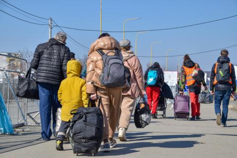 osoby widziane z tyłu idą przed siebie niosąc plecaki, torby i wózki na kółkach