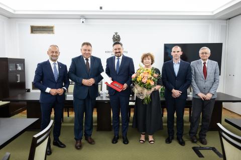 Sześć osób pozuje do zdjęcia stojąc w szeregu i uśmiechając się. W centrum stoi Marcin Wiącek trzymający w dłoniach okolicznościowy medal