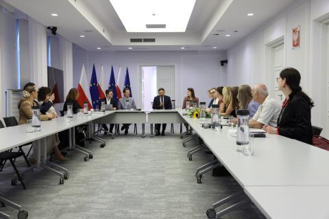 Kilkanaście osób siedzi przy stołach ustawionych w podkowę na sali konferencyjnej