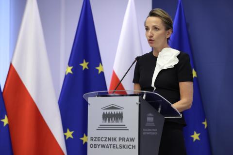 Osoba przemawia za mównicą z napisem "Rzecznik Praw Obywatelskich", w tle flagi Polski i UE