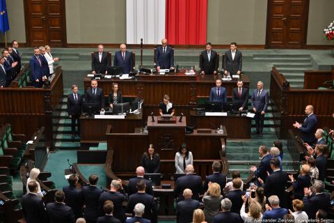 Osoba stojąca za mównicą sejmową odczytuje treść ślubowania, osoby obecne na sali obrad stoją