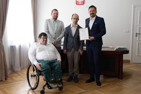 trzy osoby stoją w sali jedna siedzi na wózku 