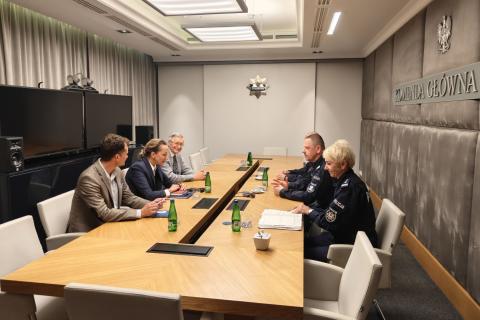 Sześć osób siedzi po dwóch stronach stołu konferencyjnego w sali z widocznym napisem "Komenda Główna Policji" na ścianie