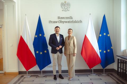 zdjęcie przedstawia dwie osoby - kobietę i mężczyznę - RPO obecnej iRPO poprzedniej kadencji pozujacych do zdjęcia na tle flag PL i UE oraz napisu Rzecznik Praw Obywatelskich