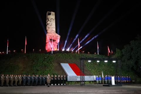 Uroczystości na Westerplatte - na kopcu stoi podświetlony biało-czerwono pomnik-obelisk otoczony masztami z flagami Polski. U stóp kopca w dwuszeregu stoi na baczność kilkadziesiąt żołnierzy.